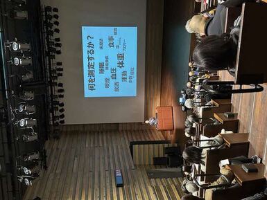 講演会の会場写真です。スライド内容は、「何を測定するか？睡眠、体重、血圧、食事、色々あります」